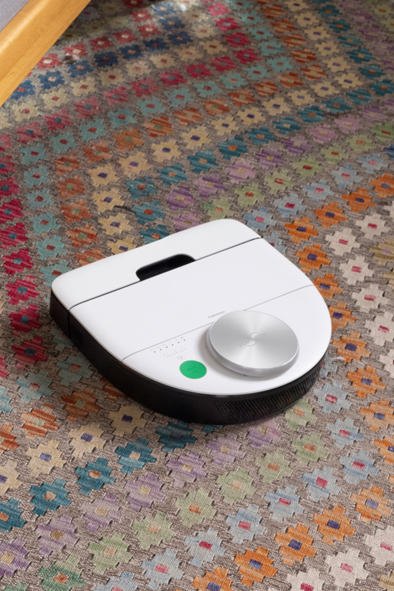 Robot aspirapolvere bianco su tappeto colorato, ripreso da vicino mentre pulisce vicino a un mobile in legno.