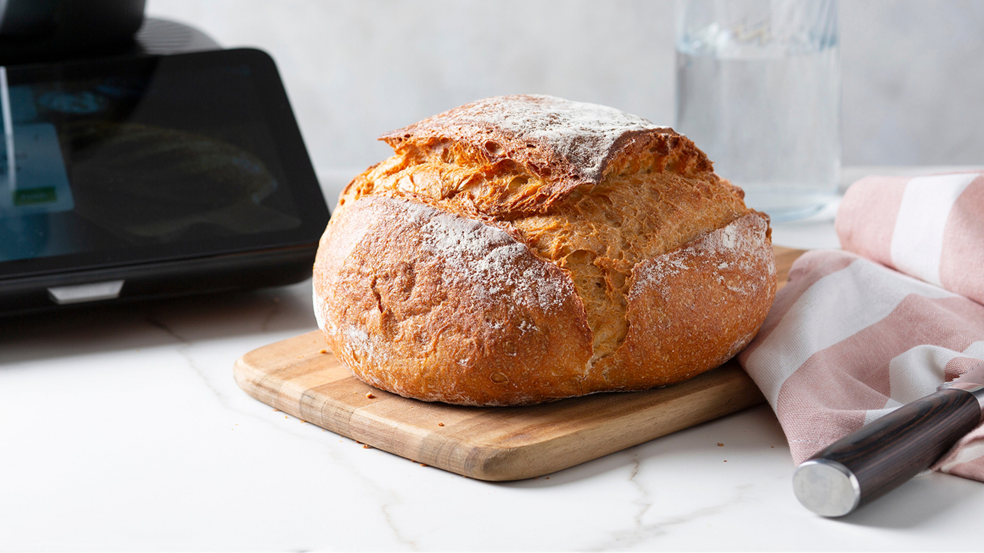 Frisch gebackenes rustikales Brot auf Holzbrett neben Thermomix® TM7, Messer, Flasche und Küchentuch.