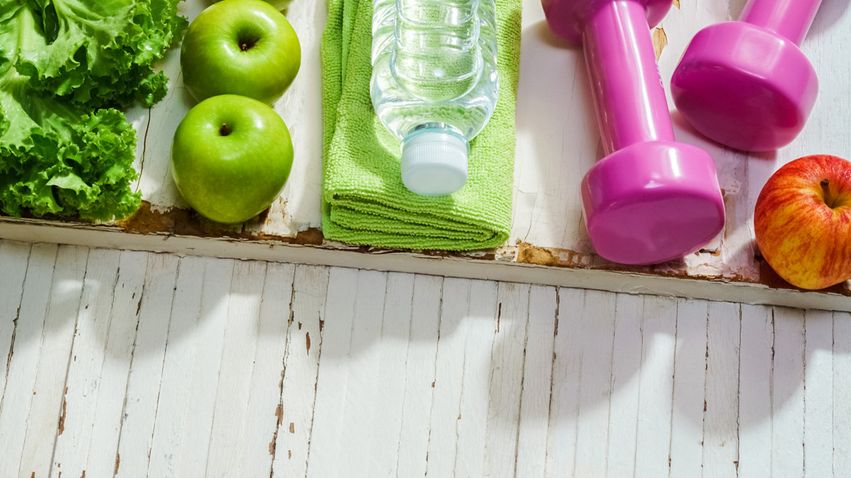 Salat, Äfpel, Wasserflasche und Hanteln auf Holztisch
