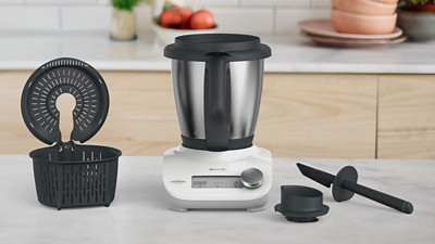 Im Bild ist eine helle und moderne Küche sichtbar. Auf der Kücheninsel im Vordergrund steht der Thermomix Friend in frontaler Ansicht. Neben dem Thermomix Friend liegt passendes Zubehör: sichtbar sind der Spatel und der Messbecher rechts vom Gerät. Links steht der Gareinsatz mit Deckel. 