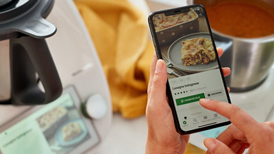 Das Bild zeigt im Hintergrund unscharf den Thermomix TM6 und den Thermomix Friend. Im Vordergrund wird ein Smartphone in den Händen gehalten. Es zeigt ein Rezept für Lasagne Bolognese auf Cookidoo – einer App mit Rezepten für den Thermomix.