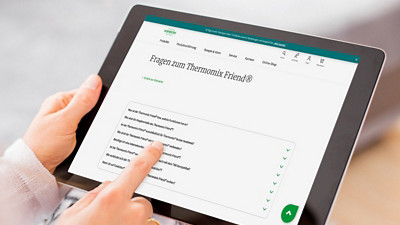 Das Foto zeigt zwei Hände, die ein schwarzes Tablet halten. Auf dem Bildschirm ist die Webseite „Fragen zum Thermomix Friend“ sichtbar. Ein Finger tippt auf den Bildschirm. 