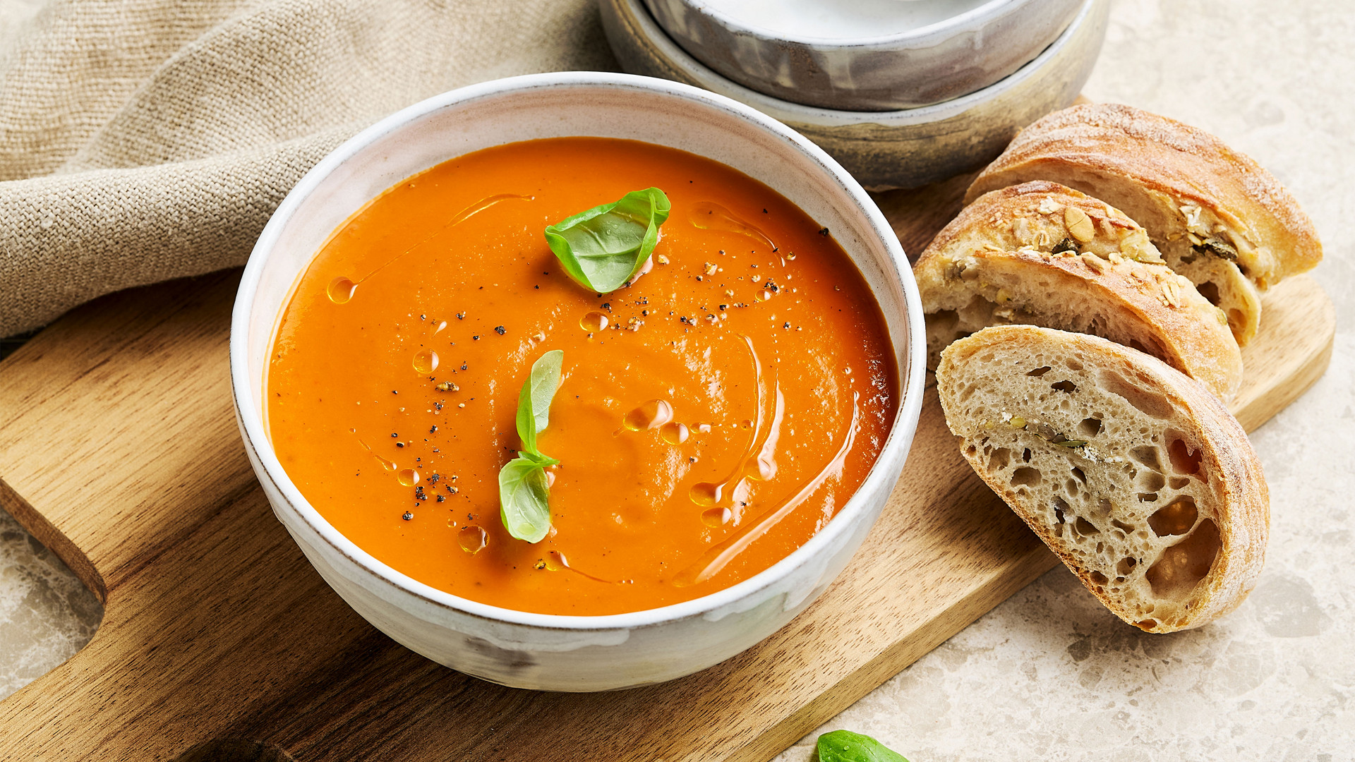 Schale cremige Tomatensuppe mit Basilikumblättern und Brotscheiben auf Holzbrett, daneben leere Schalen und Besteck.
