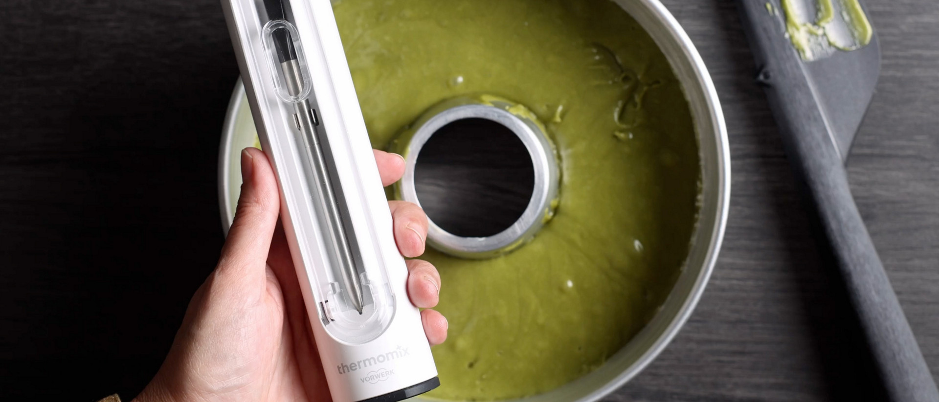 Hand hält einen Thermomix® Sensor über einer runden Backform mit grünem Teig und einem Spatel auf dunkler Oberfläche.