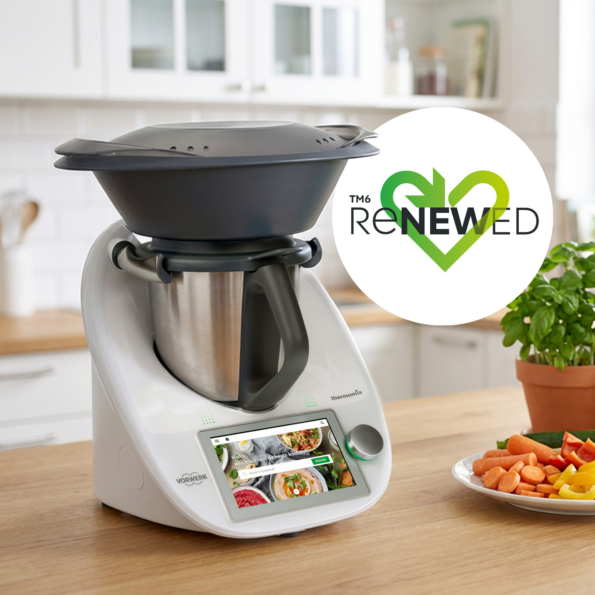 Thermomix® TM6 mit Varoma in einer Küche, "Renewed" Logo, frischem Gemüse und Kräutertöpfen auf Holztheke.