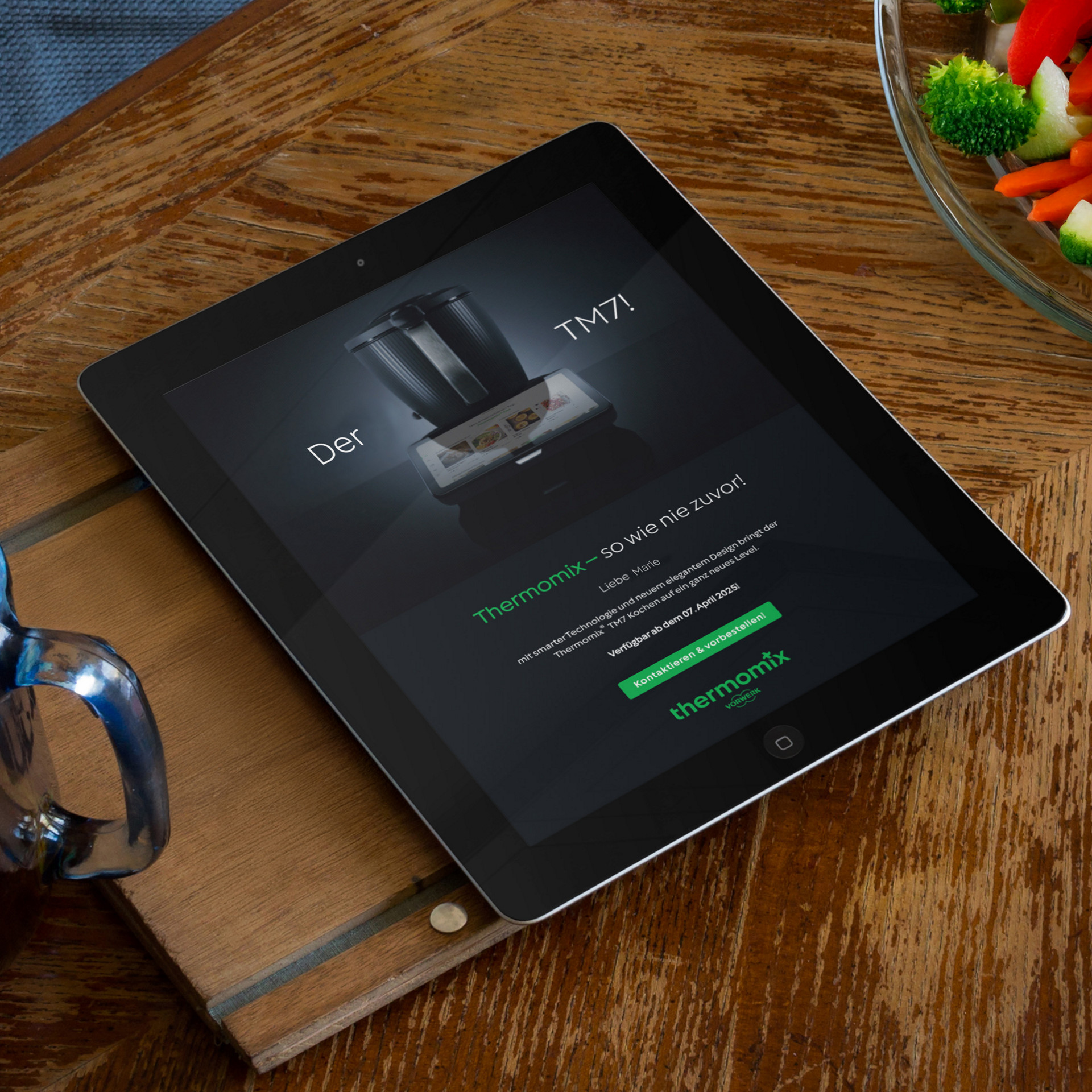 Tablet auf Holztisch zeigt eine Ankündigung für den Thermomix® TM7, daneben ein Glas und eine Schüssel mit Gemüse.