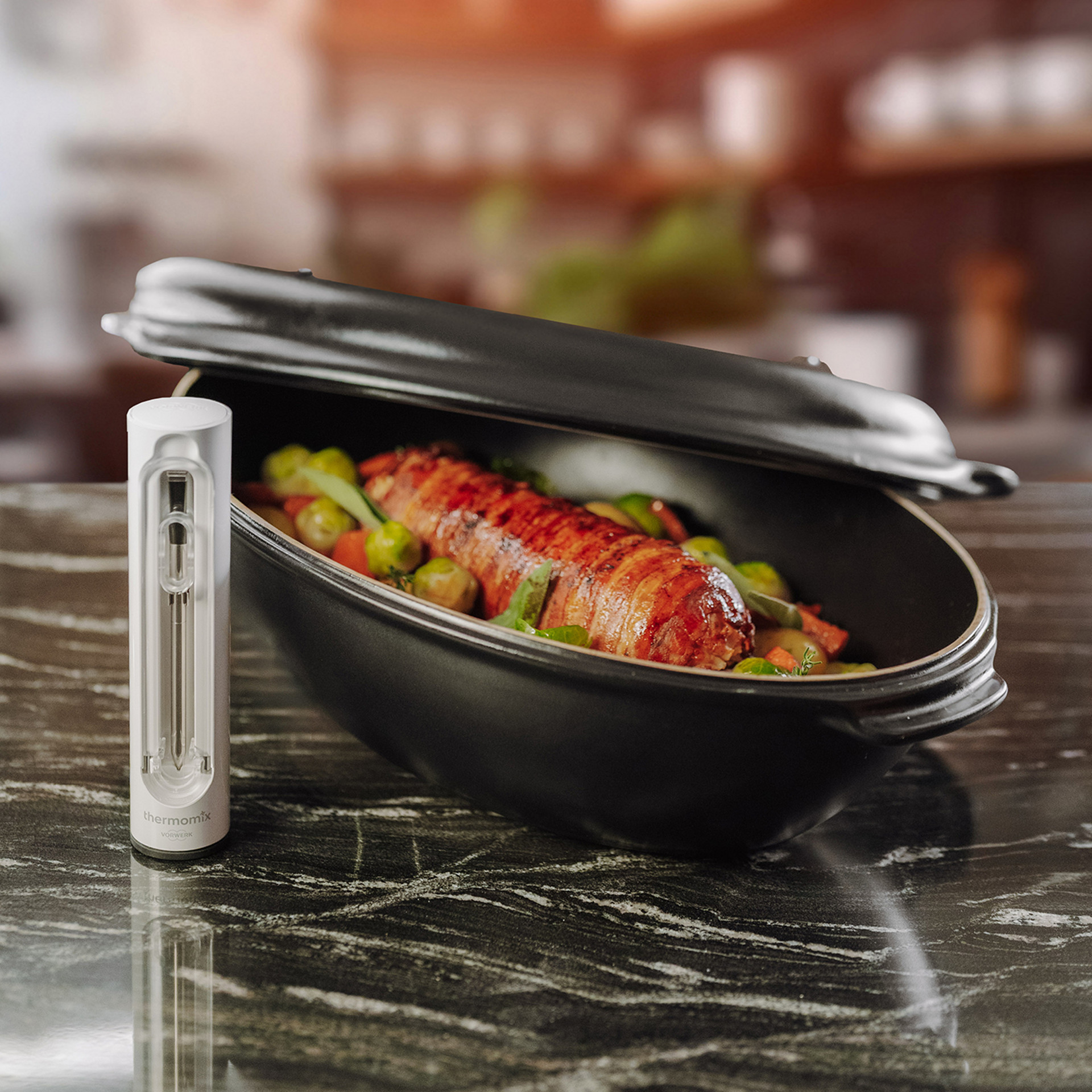 Thermomix® Sensor neben einem geschlossenen schwarzen Bräter mit Fleisch und Gemüse auf Küchenarbeitsplatte.