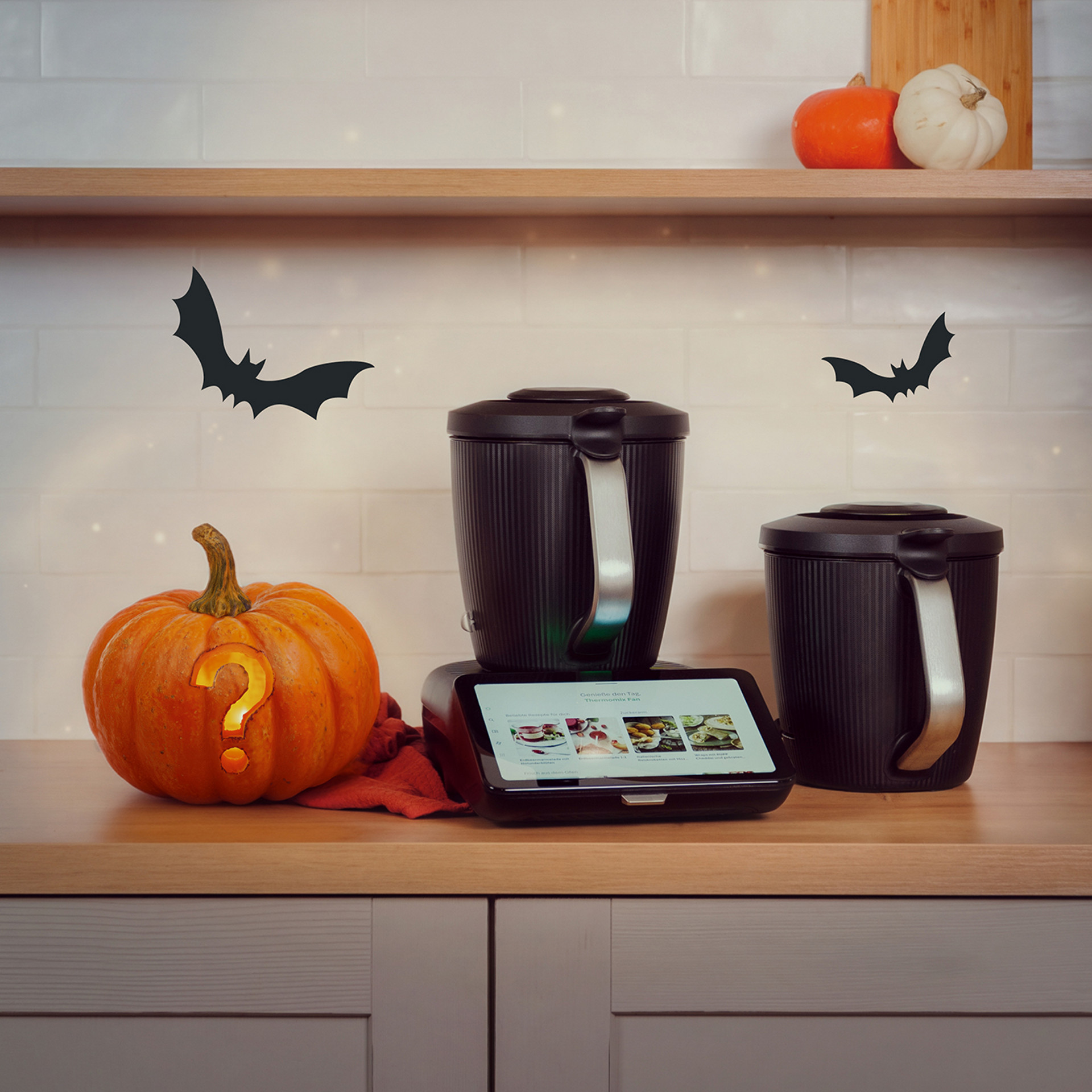 Thermomix® TM7 mit zwei Mixbehältern, Kürbis mit Fragezeichen und Halloween-Fledermäusen auf einer Küchenarbeitsplatte.