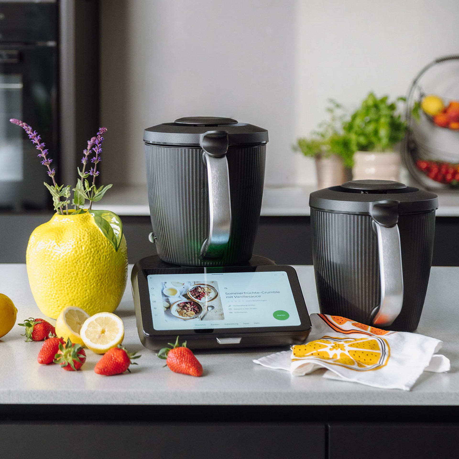 Thermomix® TM7 mit Mixbehältern auf einer Küchenarbeitsplatte, umgeben von Zitronen, Erdbeeren und einer Vase mit Kräutern.