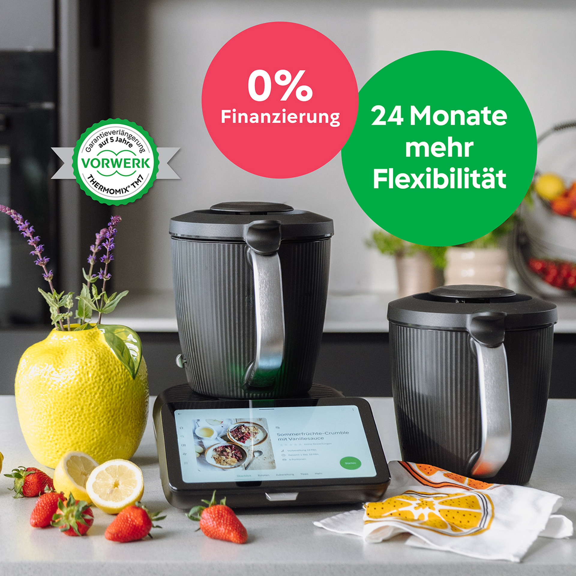 Thermomix® TM7 mit zwei Mixtöpfen auf der Arbeitsplatte, Früchte und Tuch daneben, Finanzierungsangebot im Hintergrund.