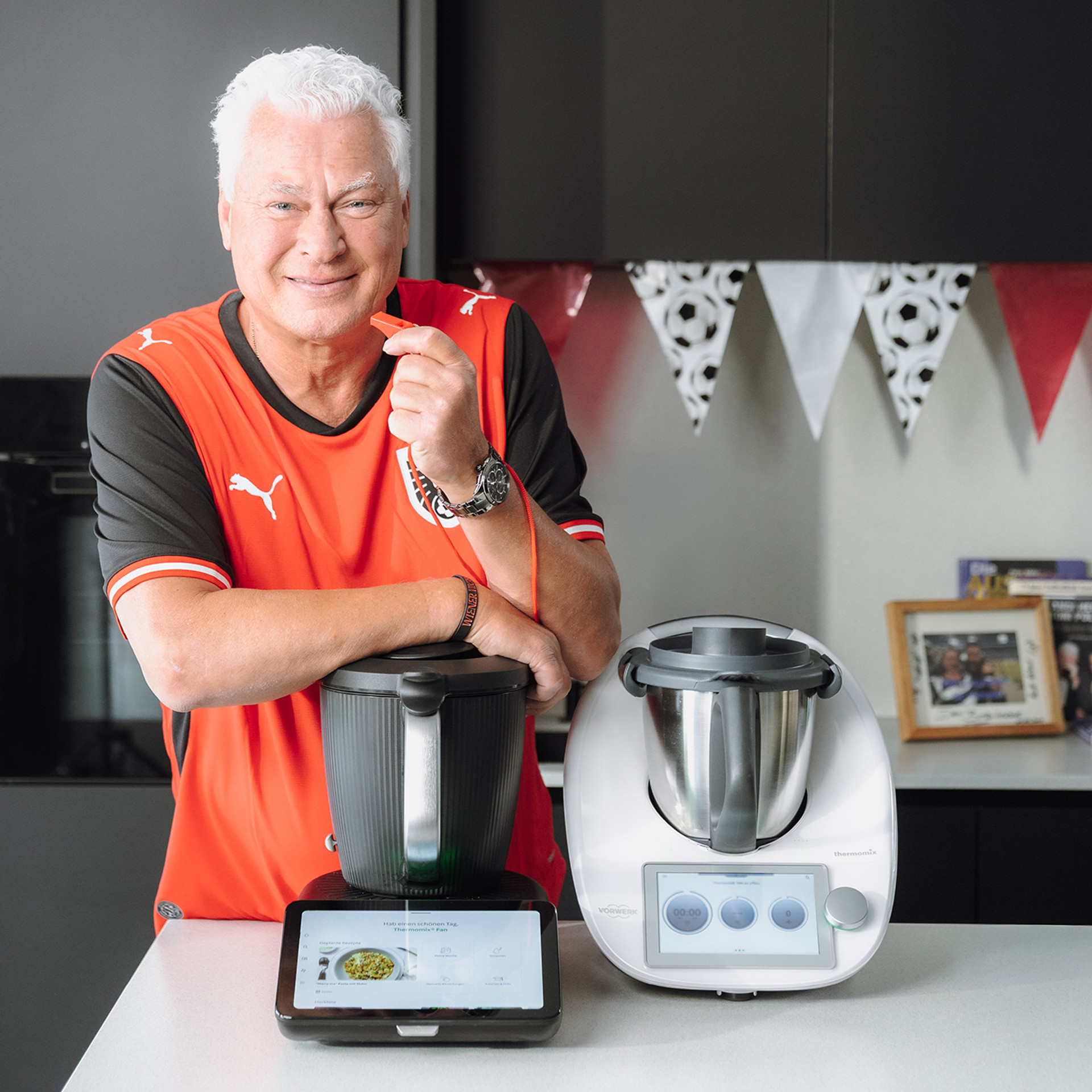 Toni Polster mit rotem Sportshirt mit Thermomix® TM7 und Thermomix® TM6 auf einer Küchenarbeitsplatte.