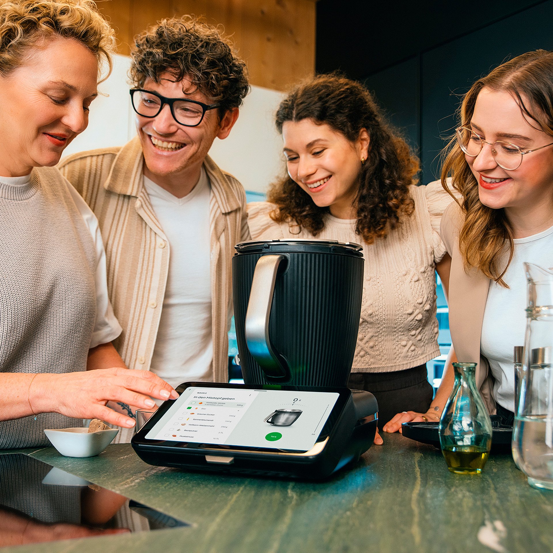 Vier Personen in einer Küche bereiten mit dem Thermomix® TM7 auf der Arbeitsplatte Speisen zu und folgen dem Rezept auf dem Display.