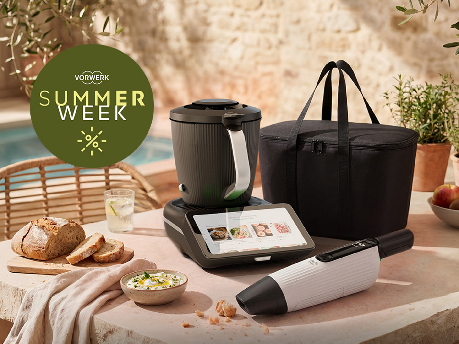 Thermomix® TM7 und Kobold VM7 auf einem Tisch mit Brot, Dip, Obst und schwarzer Tasche während Vorwerk Summer Week.