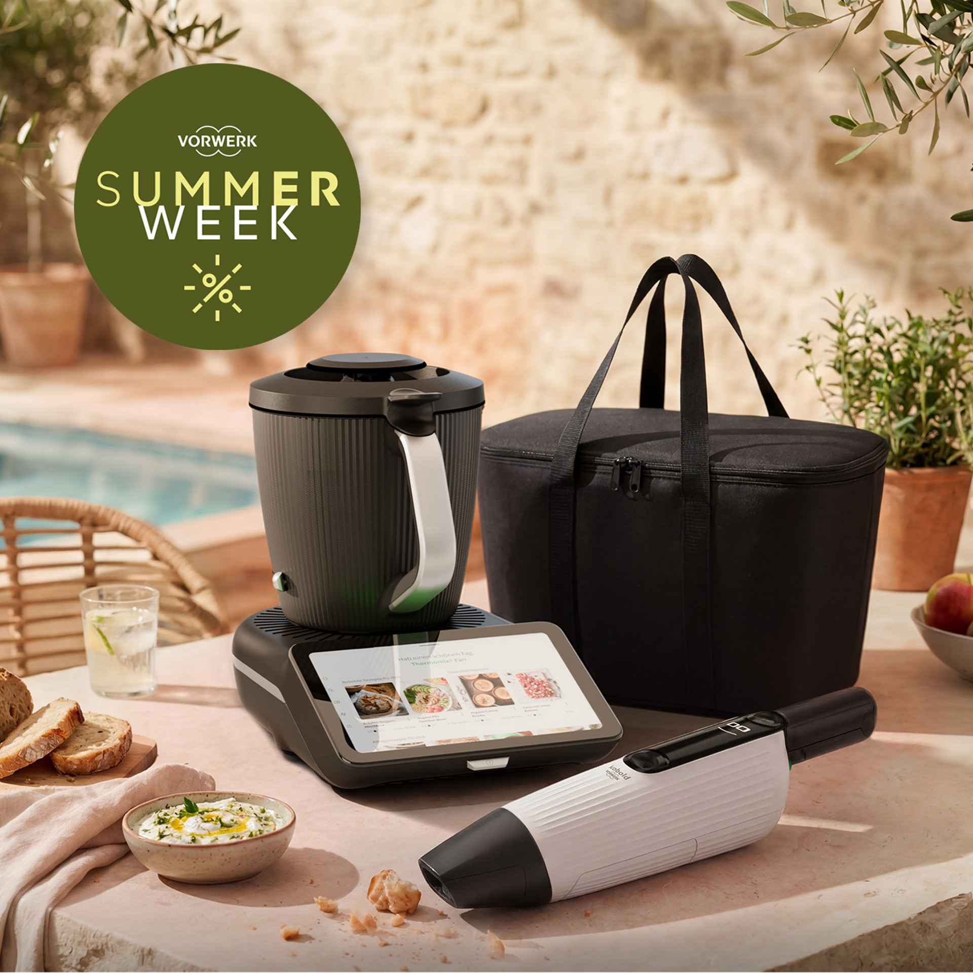 Thermomix® TM7 und Kobold VM7 auf einem Tisch mit schwarzer Tragetasche, Brot und Dip in sommerlicher Umgebung.