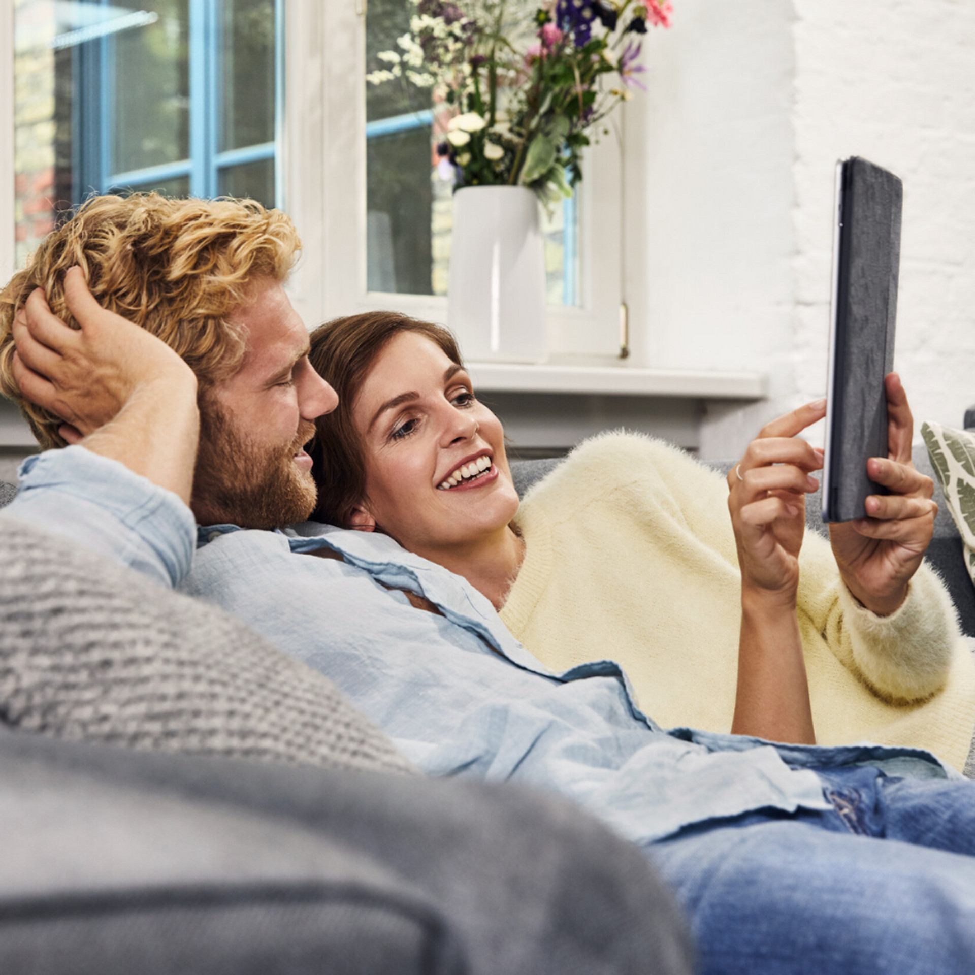 Paar entspannt auf einem Sofa und schaut auf ein Tablet, im Hintergrund eine Vase mit Blumen auf dem Fensterbrett.
