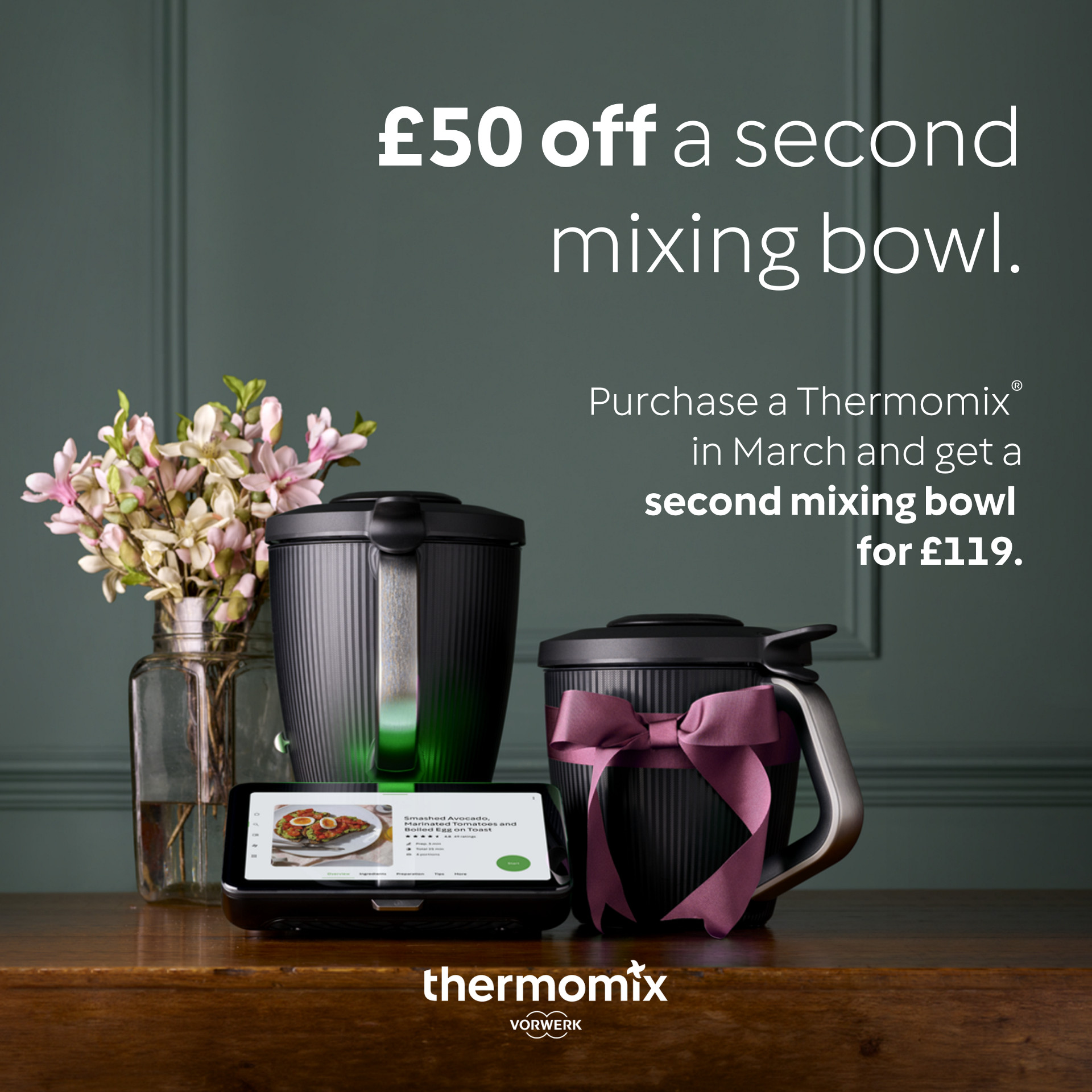 Thermomix® TM7 with touchscreen, a second mixing bowl with a purple ribbon, and flowers on a green table, £50 off offer.
