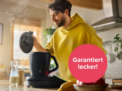 Ein Mann hebt lächelnd in der Küche den Deckel des dampfenden Thermomix® TM7, während Zutaten wie Milch und Getreide bereitstehen.