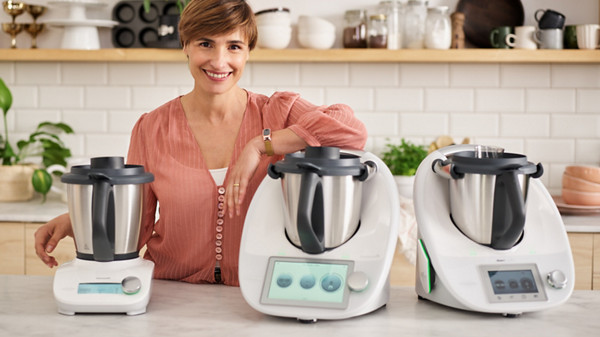 Im Bild ist eine helle und moderne Küche abgebildet. Auf der Arbeitsplatte der Kücheninsel stehen der Thermomix TM6, der Thermomix TM5 und der Thermomix Friend. Eine Frau mit kurzen, bräunlichen Haaren und einer rosafarbenen Bluse lehnt mit einem Arm lächelnd auf dem Thermomix TM6.