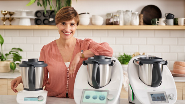 Im Bild ist eine helle und moderne Küche abgebildet. Auf der Arbeitsplatte der Kücheninsel stehen der Thermomix TM6, der Thermomix TM5 und der Thermomix Friend. Eine Frau mit kurzen, bräunlichen Haaren und einer rosafarbenen Bluse lehnt mit einem Arm lächelnd auf dem Thermomix TM6.