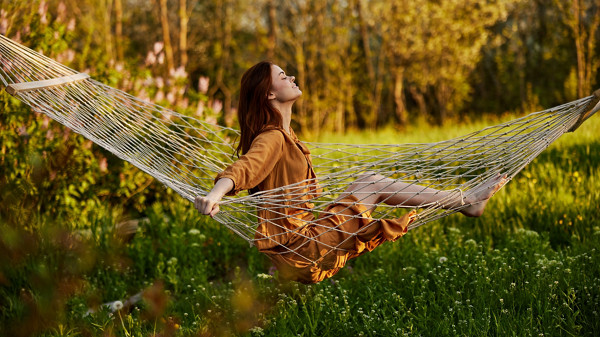 uma mulher feliz, com um longo vestido cor de laranja, descansa sentada numa rede na dacha, sorrindo agradavelmente, olhando para longe, iluminada pelo sol de verão durante o pôr do sol. Fotografia horizontal