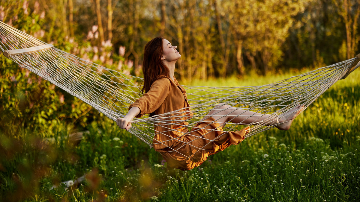 uma mulher feliz, com um longo vestido cor de laranja, descansa sentada numa rede na dacha, sorrindo agradavelmente, olhando para longe, iluminada pelo sol de verão durante o pôr do sol. Fotografia horizontal