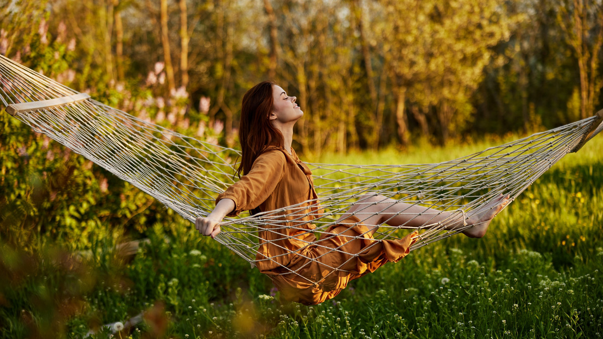 a happy woman in a long orange dress is resting sitting in a hammock at the dacha, smiling pleasantly looking away, illuminated by the summer sun during sunset. Horizontal photo