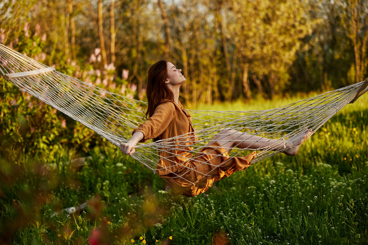 uma mulher feliz, com um longo vestido cor de laranja, descansa sentada numa rede na dacha, sorrindo agradavelmente, olhando para longe, iluminada pelo sol de verão durante o pôr do sol. Fotografia horizontal