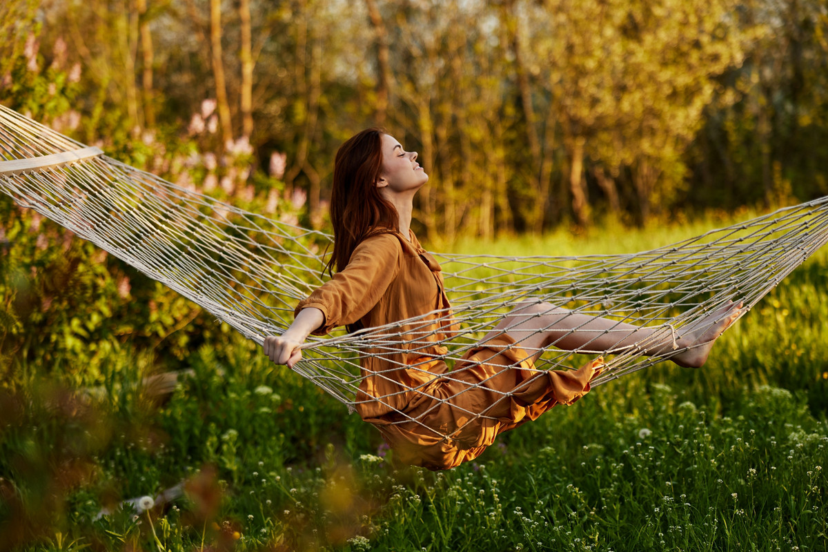 a happy woman in a long orange dress is resting sitting in a hammock at the dacha, smiling pleasantly looking away, illuminated by the summer sun during sunset. Horizontal photo