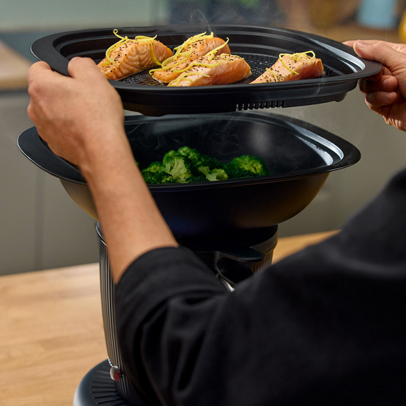 Person placing salmon in steaming basket on Thermomix TM7 with broccoli in Varoma in a kitchen setting