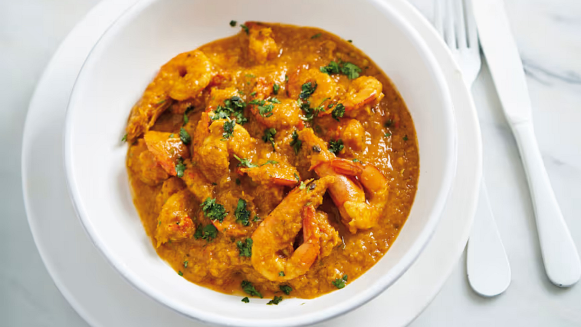 Tigela branca com caril de camarão guarnecido com ervas, sobre prato e talheres brancos.