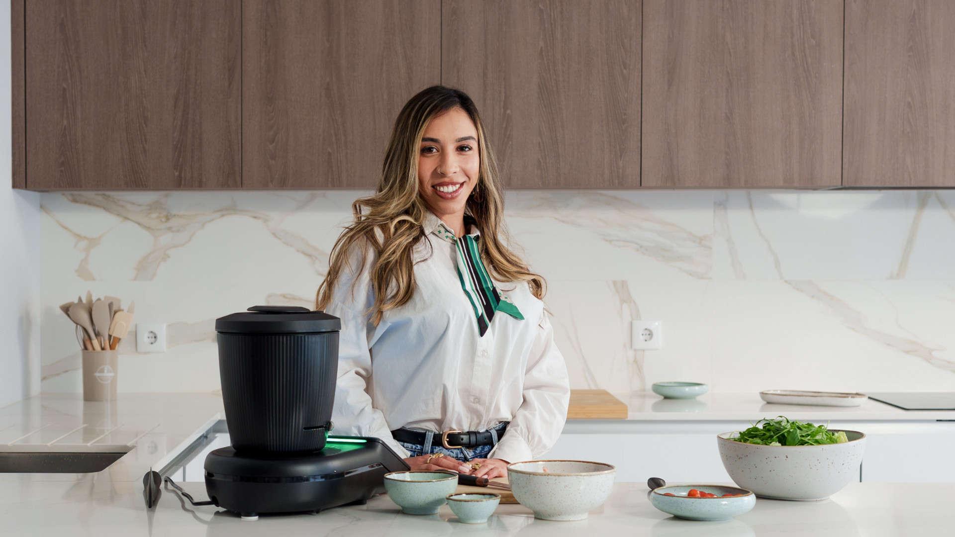 Mulher numa cozinha moderna ao lado de um Bimby® TM7 com taças de legumes no balcão.