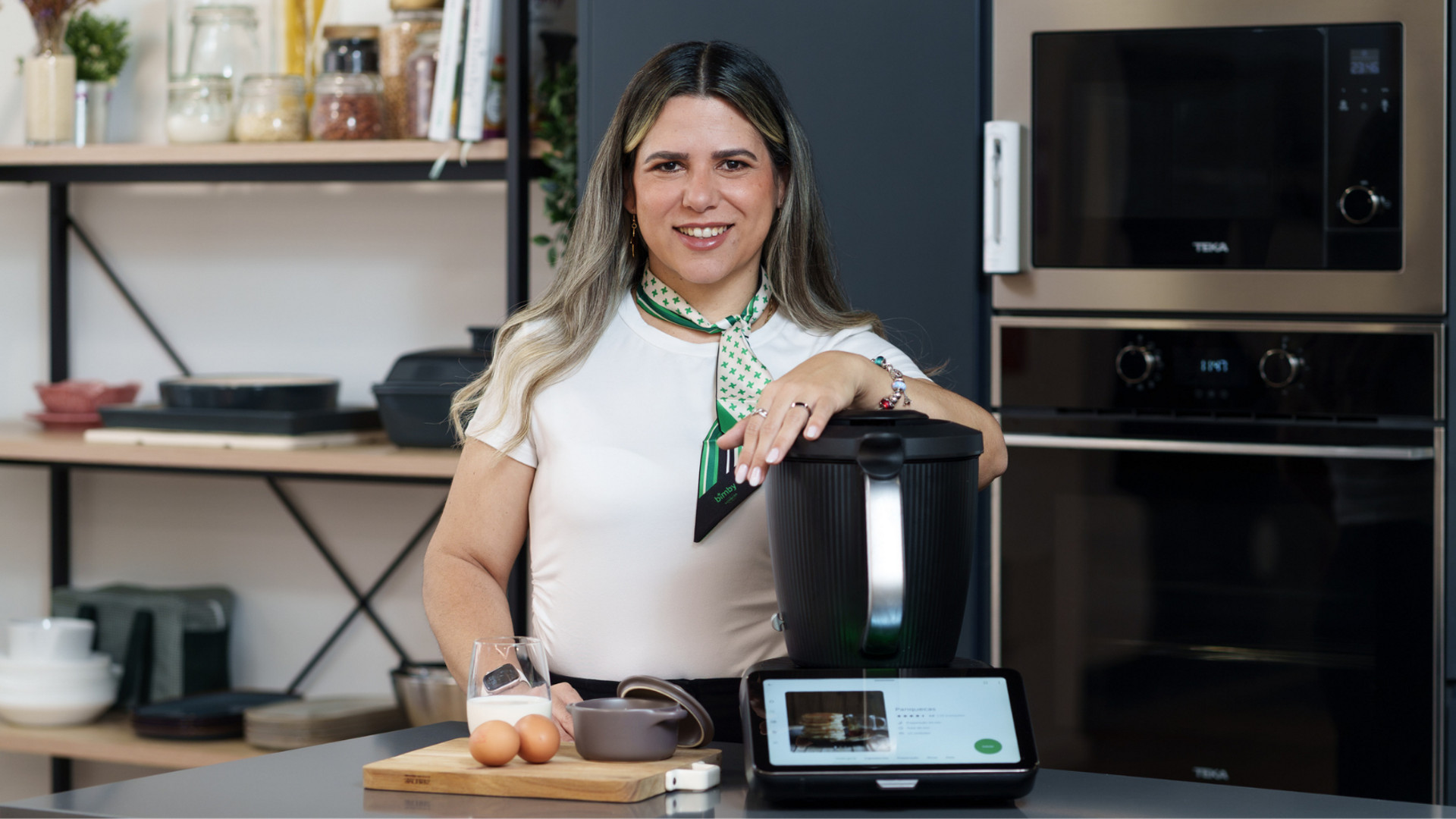 Mulher numa cozinha com Bimby® TM7, ovos, jarro de leite e utensílios numa tábua de cortar.