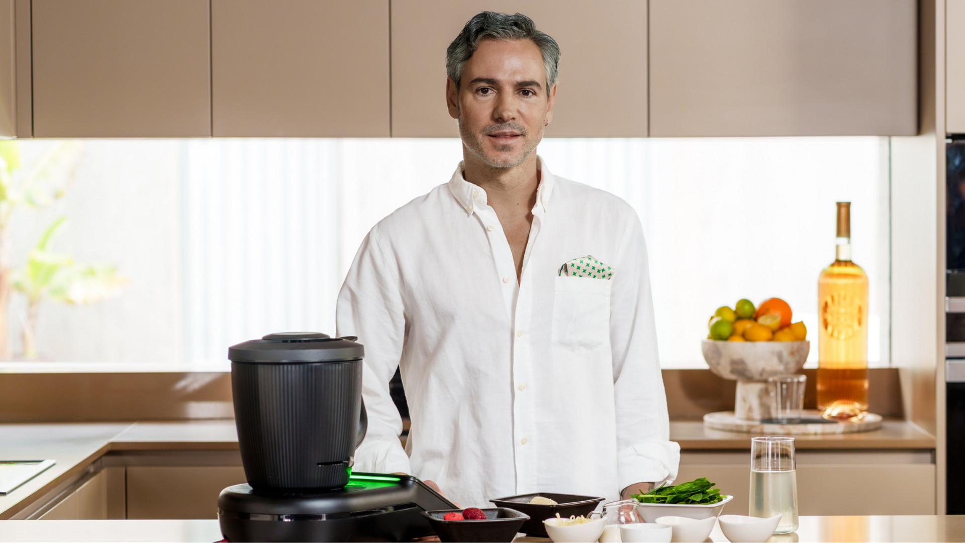 Homem na cozinha atrás de um Bimby® TM7 com vários ingredientes em taças no balcão.