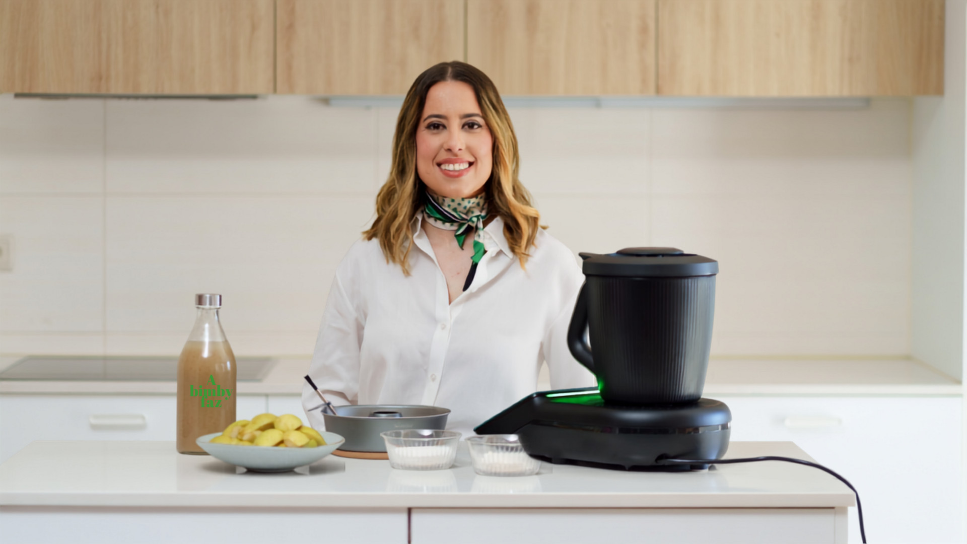 Mulher na cozinha sorrindo atrás do Bimby® TM7, com uma taça de batatas, garrafa personalizada e ingredientes no balcão.