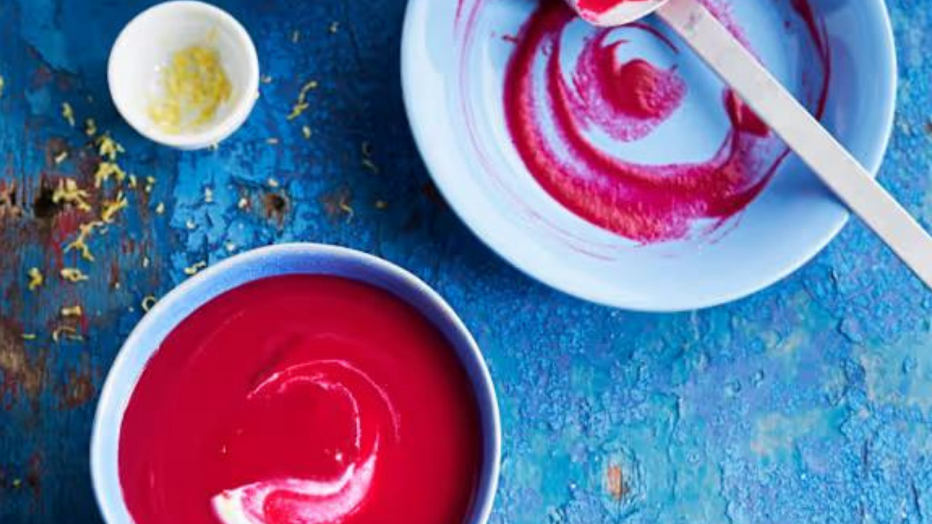 Tigela com sopa cor-de-rosa, tigela vazia com resíduos vermelhos e colher, e pequena tigela com raspas de limão sobre fundo azul.