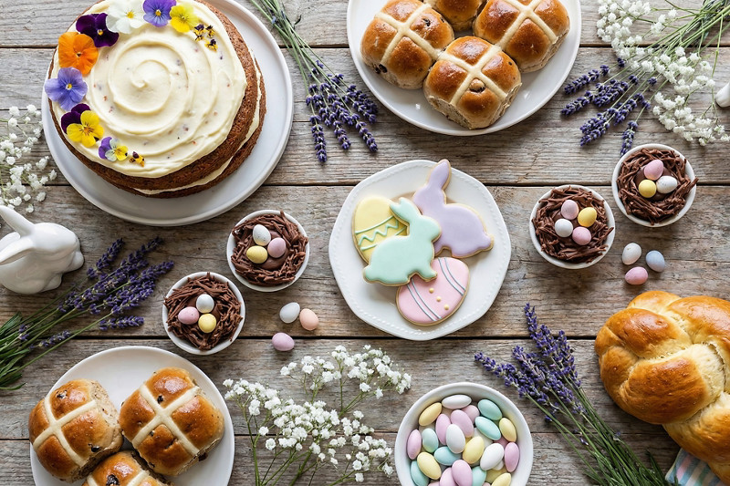 Easter celebration food spread featuring hot cross buns, decorated cake, bunny and egg cookies, chocolate nest cupcakes, lemon bars, and candy eggs on a rustic wooden table