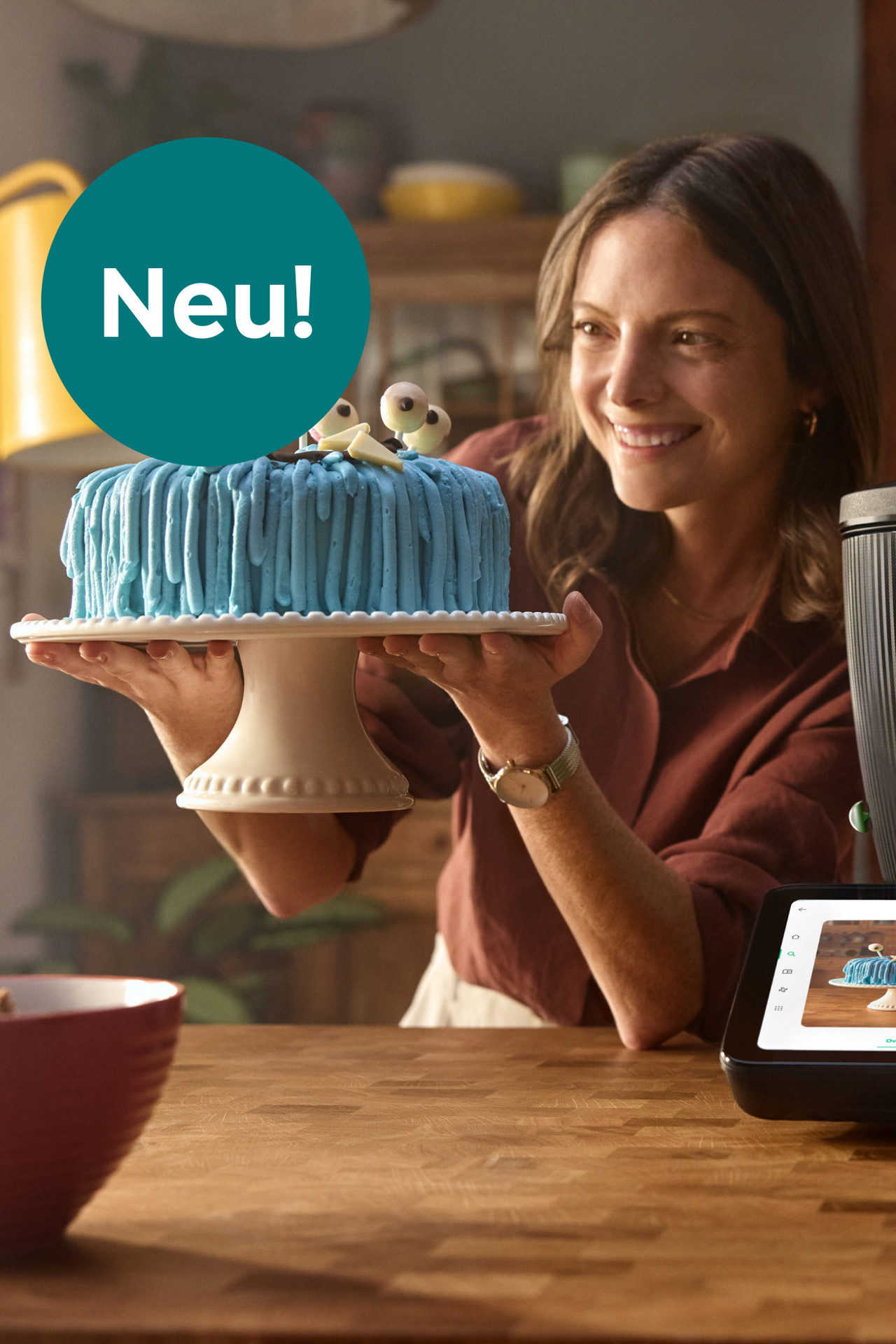 Person hält eine Torte mit blauer Glasur und Augen, neben Thermomix® TM7; Kreis mit Schriftzug „Neu!“.