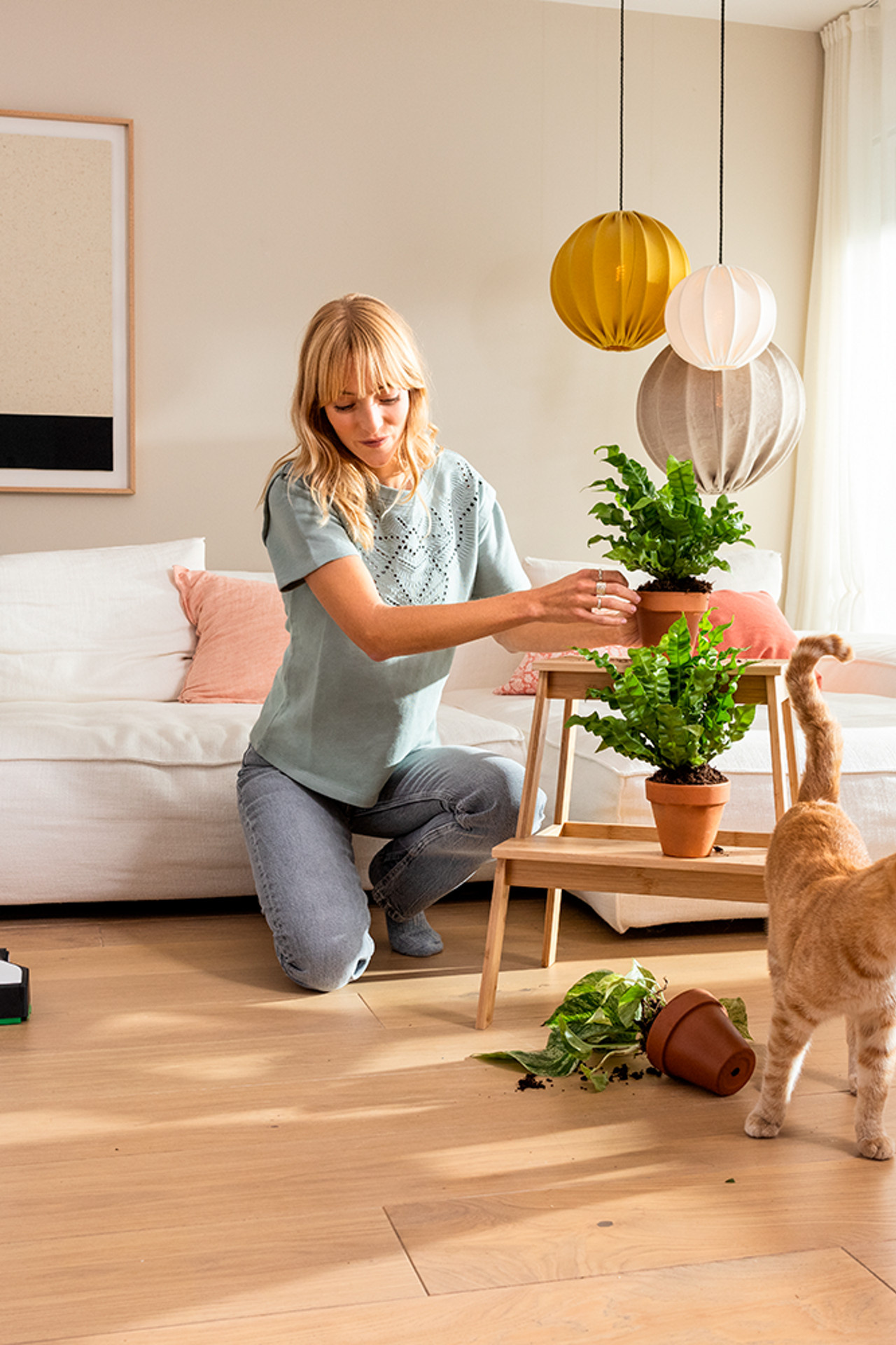 Kobold VK7 está sobre el suelo de madera, una mujer organiza plantas y un gato naranja camina cerca.
