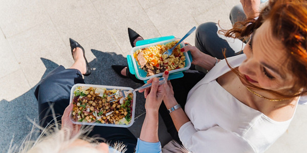 Capturing a top view of three businesswomen sitting together outdoors, enjoying healthy meals from lunch boxes during their break, fostering communication and friendship in a vibrant urban setting