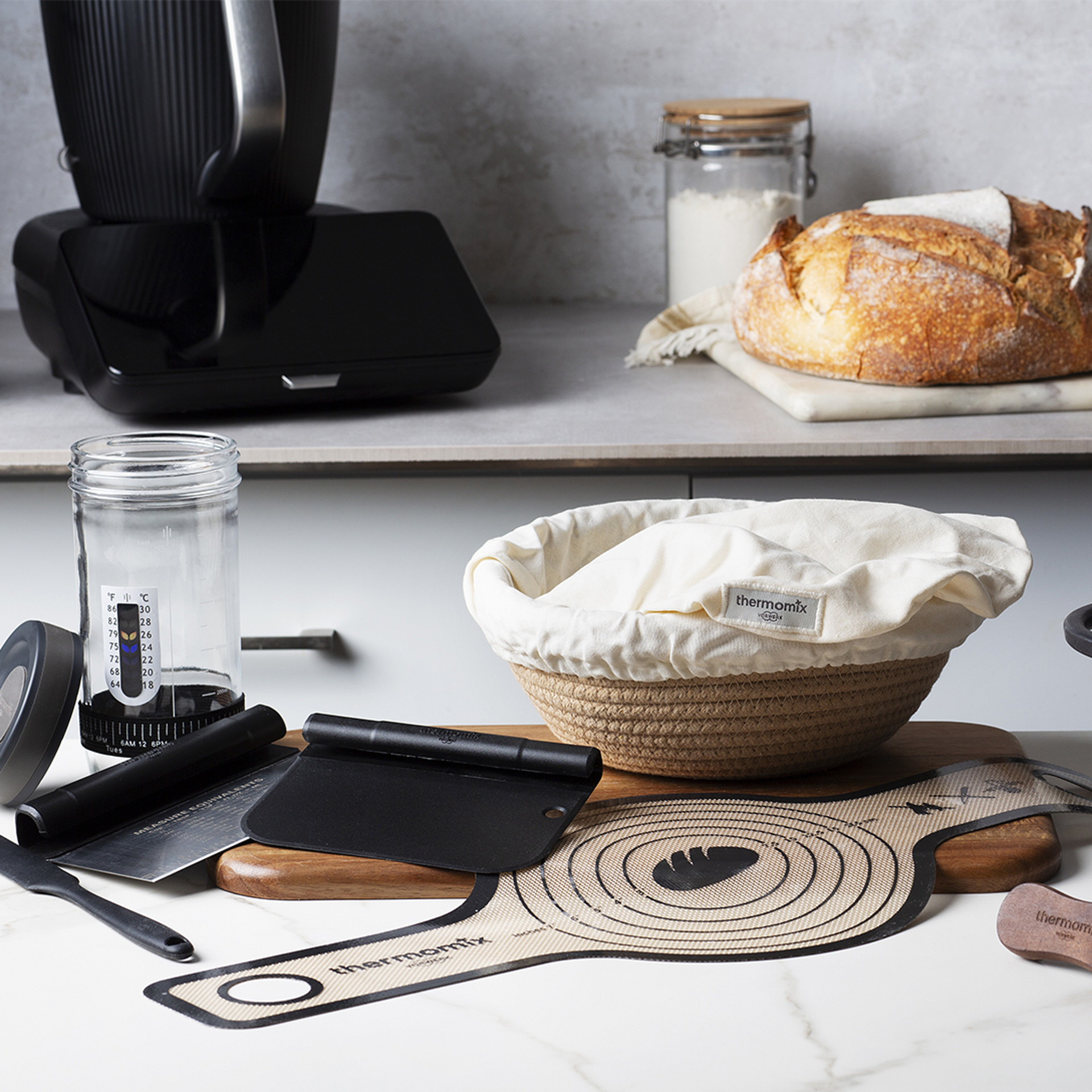 Cuisine avec Thermomix® TM7, pain, panier de pousse, bocal, coupe-pâte et tapis de cuisson sur le plan.