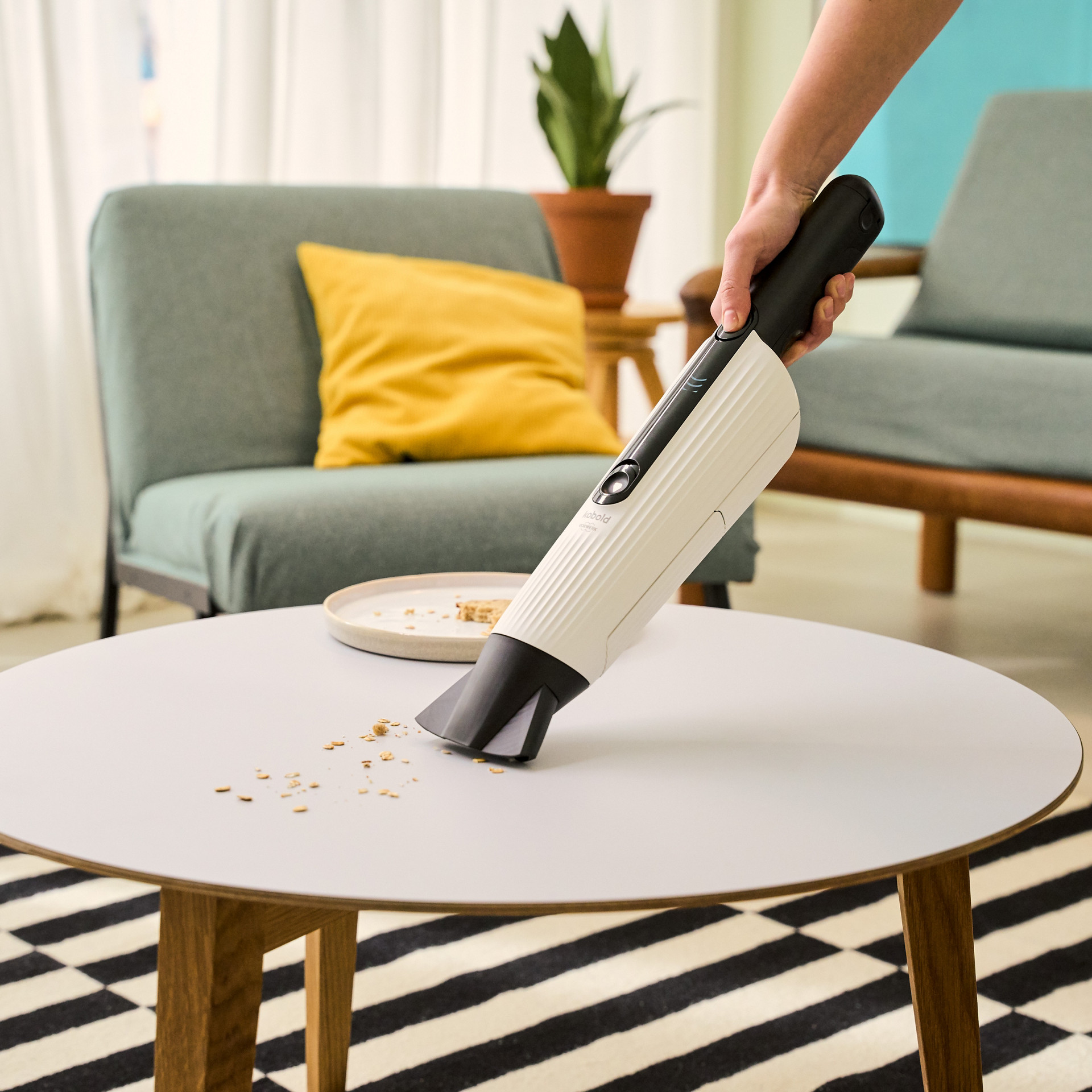 Personne nettoyant des miettes sur une table basse ronde avec l’aspirateur de table Kobold VM7 dans un salon.
