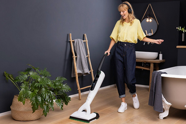 Une femme passe l'aspirateur-laveur Kobold VK7 dans sa salle de bain en écoutant de la musique