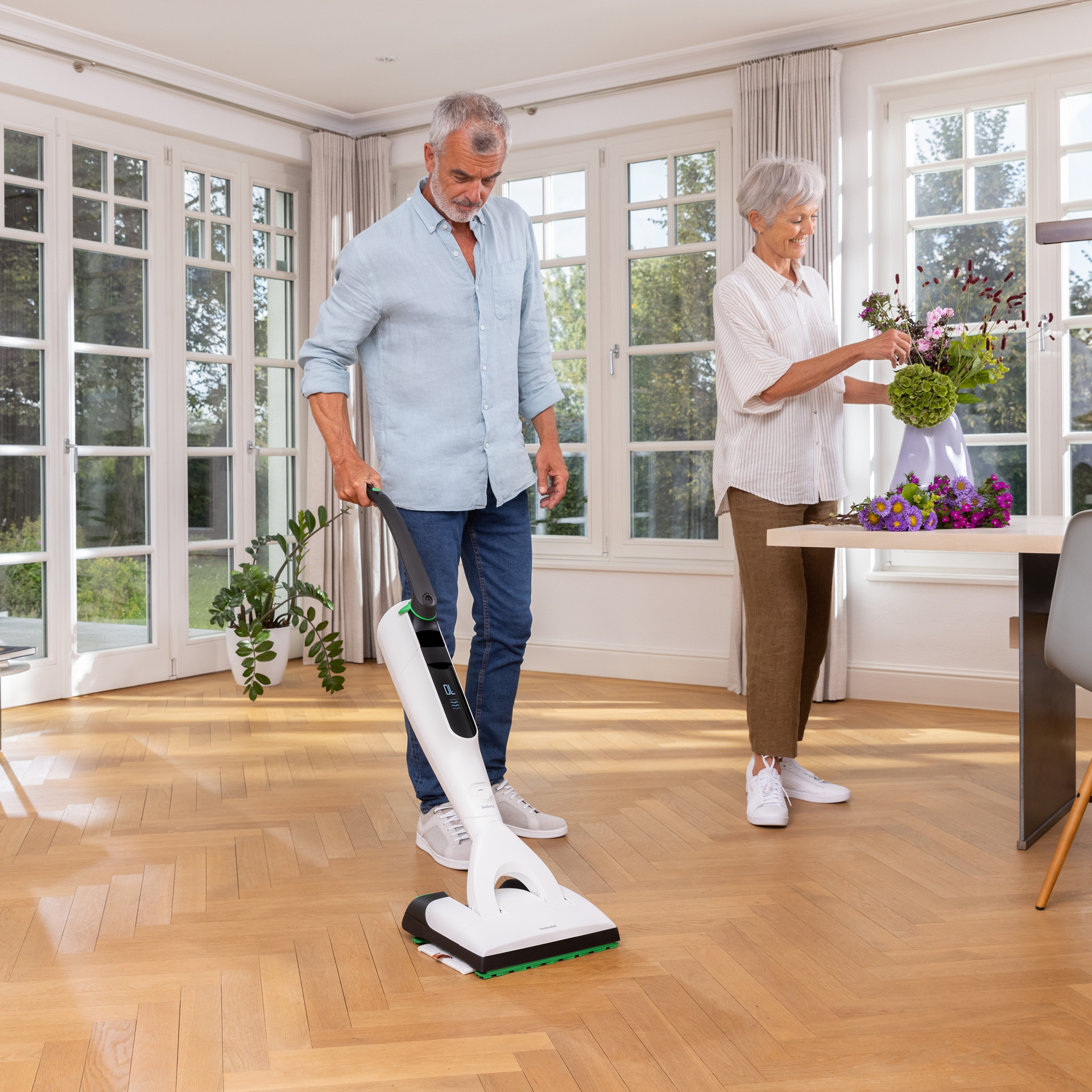 Homme utilisant l’aspirateur laveur sans fil Kobold VK7 avec brosse DuoSpeed SP7 dans un salon lumineux, femme arrange des fleurs.