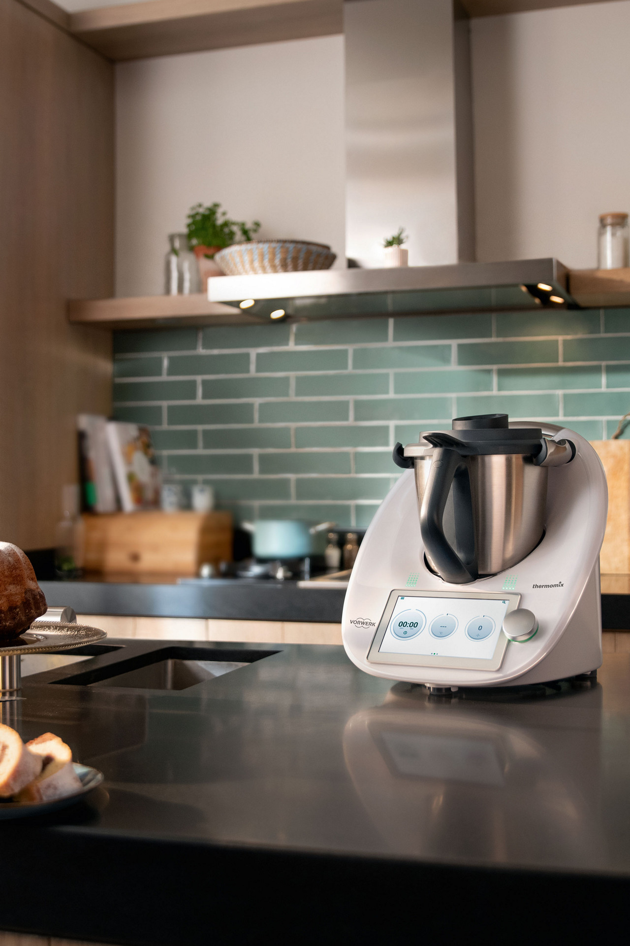 Thermomix® TM6 sur un plan de travail de cuisine avec des ingrédients de pâtisserie et un gâteau marbré au premier plan.