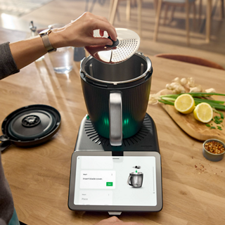 Person placing Thermomix Cutter Plus blade into Thermomix TM7 on a kitchen counter with ingredients nearby