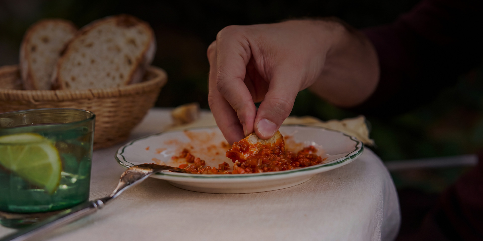 Hand tunkt Brot in einen Teller mit Tomatensauce, daneben Brotkorb und Getränk mit Limetten auf dem Tisch.