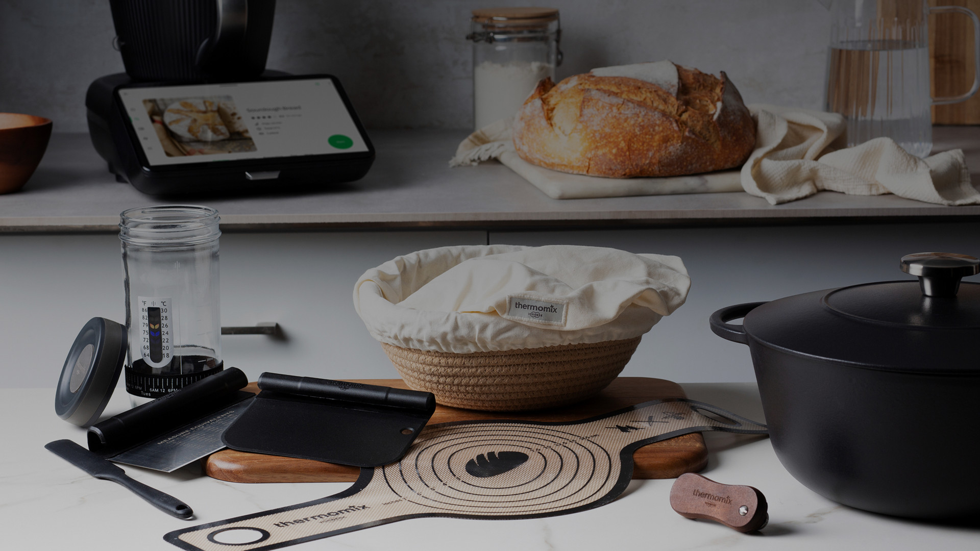 Thermomix® TM7 mit Brotbackzubehör auf der Arbeitsfläche, inklusive Gärkorb und Brotlaib.