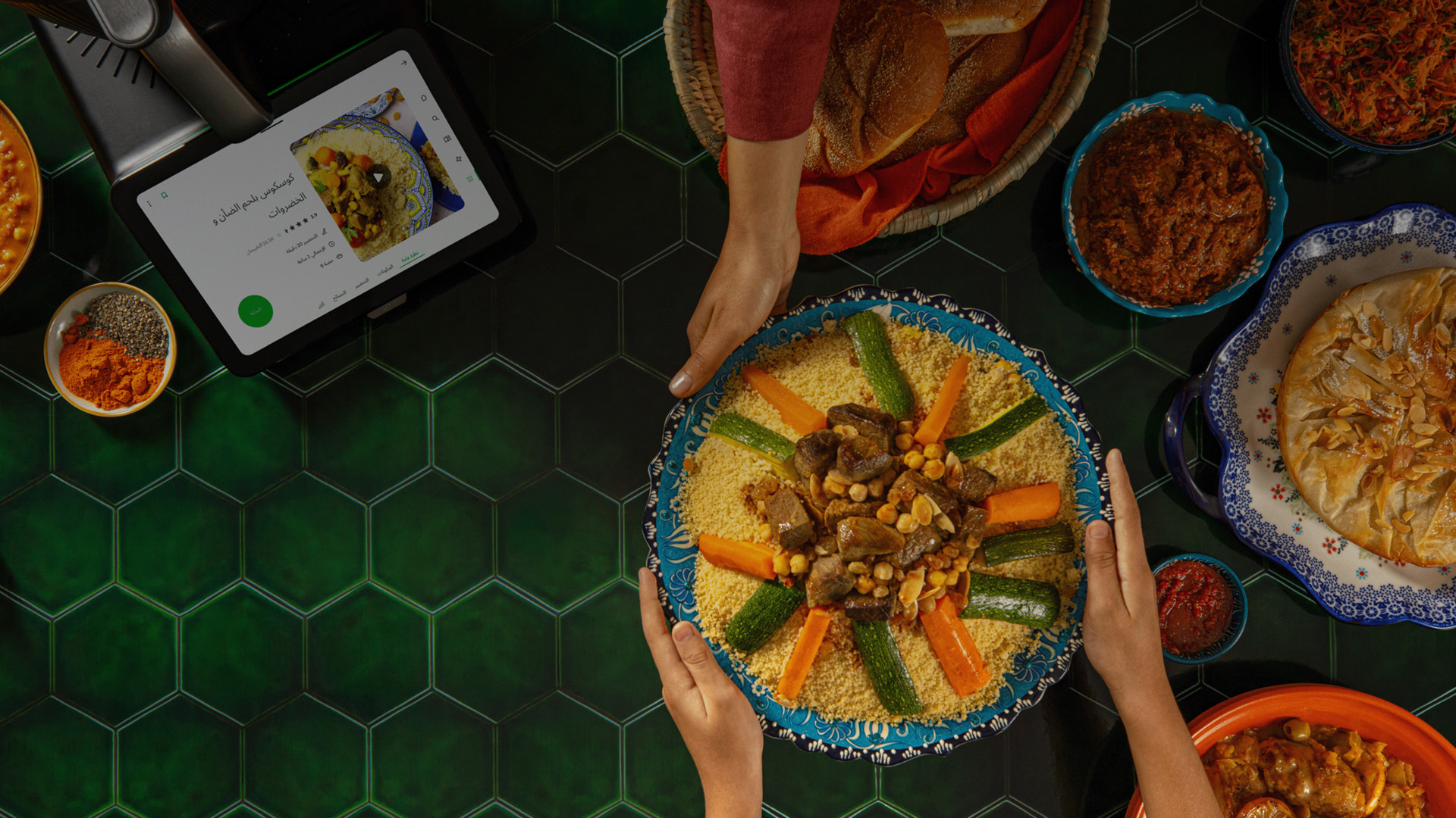 Three hands sharing a large bowl of couscous with vegetables and lamb, Thermomix® TM6 displaying a recipe, surrounded by Moroccan dishes.