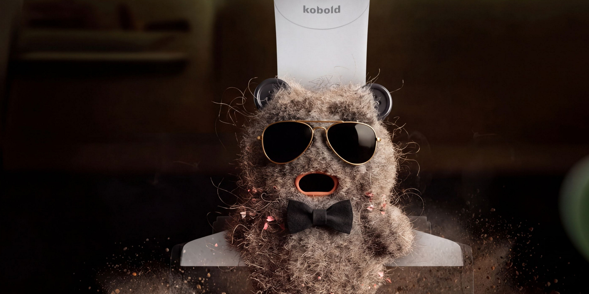 Kobold VK7 Saugerkopf nimmt eine Staubflocke mit Sonnenbrille und Fliege auf, umgeben von Staub und Schmutz.