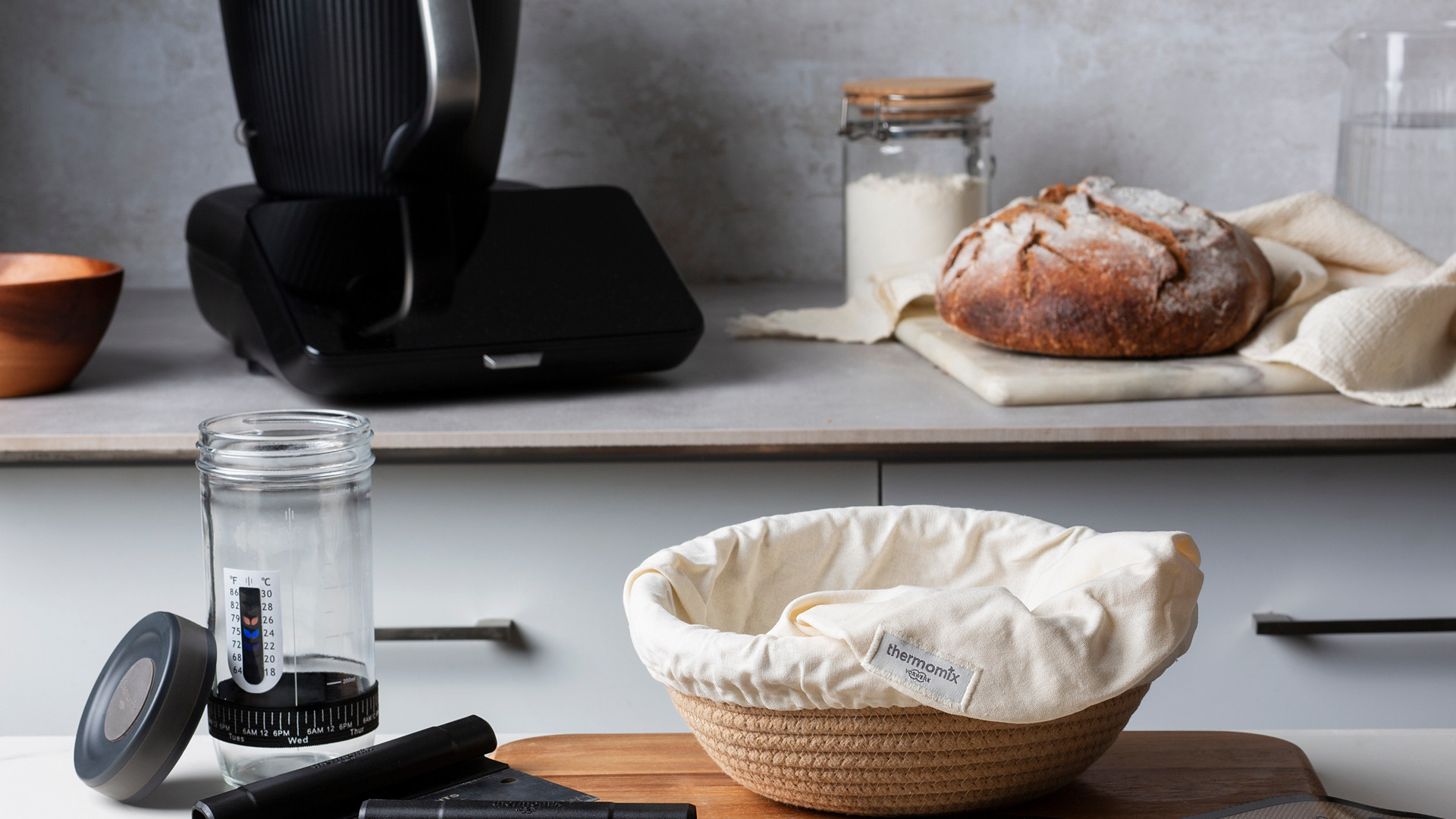 Thermomix® TM7 auf einer Küchenarbeitsplatte mit Brotbackzubehör, Gärkörbchen, Brot, Mehldose und Wasserkrug.